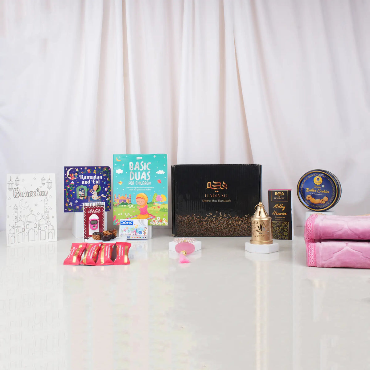 A front-facing arrangement of the Ultimate Kids Celebration Hamper contents, featuring a black Hadiyah gift box, a folded pink velvet prayer mat, children's Islamic books, a coloring sheet, a cookie tin, chocolates, a pink tasbeeh, and a mini golden lantern.