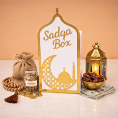 White Sadaqah Box with gold detailing, surrounded by coins, dates, and lantern, ideal for housewarming and Ramadan gifts in Religious Items.
