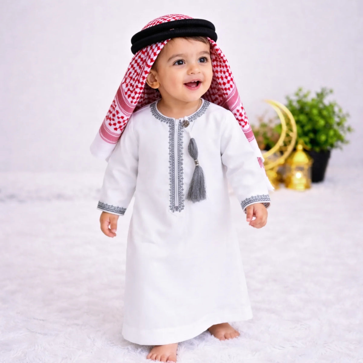 Baby boy wearing traditional white thobe with embroidery, standing on white fluffy rug