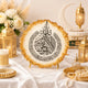 A round decorative plate with Arabic calligraphy in black on a white background, framed by a glittering gold scalloped edge, displayed on a draped white fabric table with ornate gold lanterns, candles, flowers, and a crescent moon decor in the background.