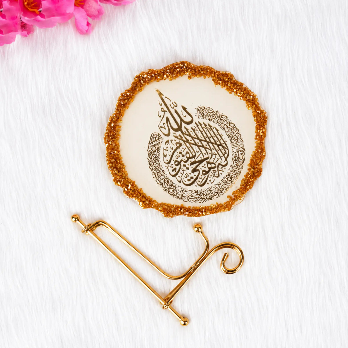 A round decorative plaque with Arabic calligraphy in dark brown on an off-white center, surrounded by a glittery gold border, displayed on a gold stand against a white textured background with pink flowers in the corner.
