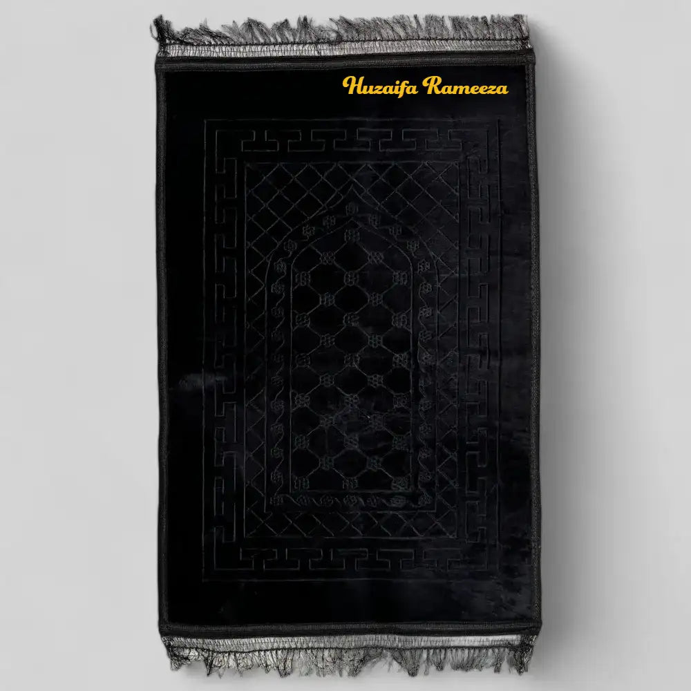 Black prayer mat with intricate design and 'Huzaija Rameeza' branding on a light gray background