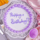 Top view of Floral Vanilla Cream Cake featuring purple floral accents and 'Happy Birthday' icing text, perfect for elegant celebrations.