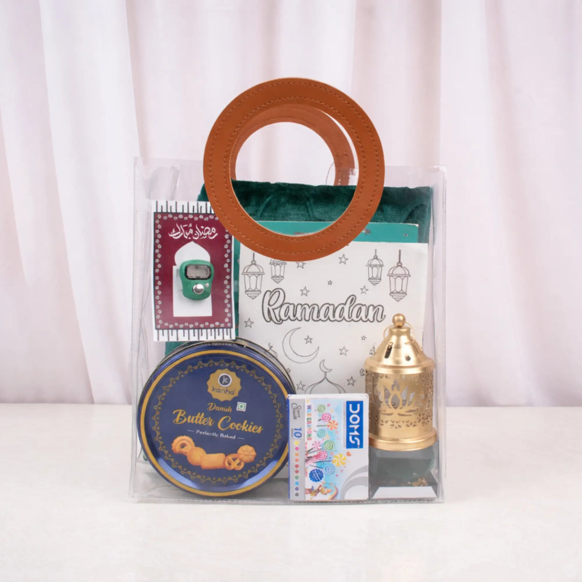 The green-themed Imaan Play & Pray Kids Hamper fully assembled inside a clear PVC bag with round brown handles. A green prayer mat, Basic Duas book, green digital tasbeeh, golden lantern, cookie tin, and crayons are visibly neatly packed inside.