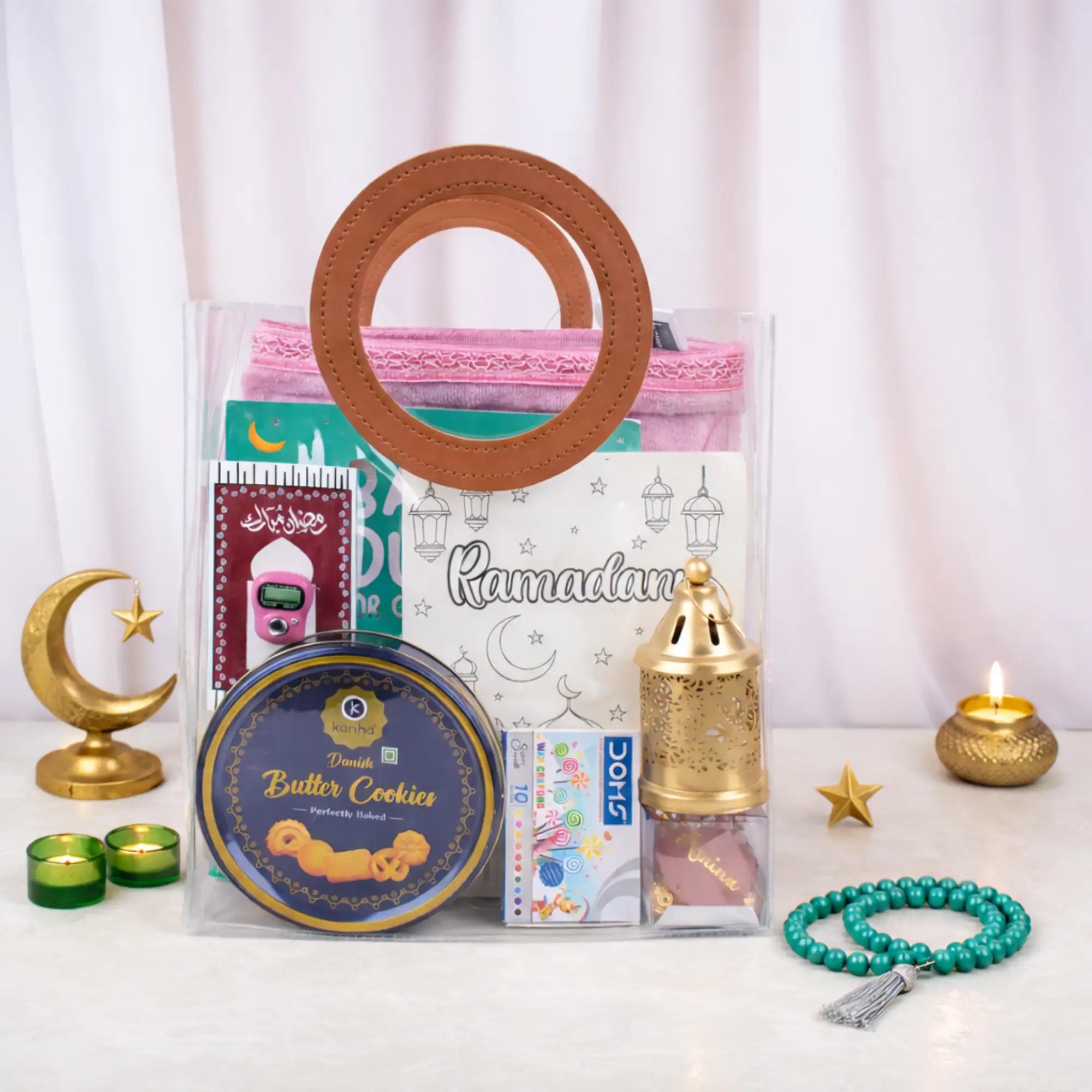 The pink-themed Imaan Play & Pray Kids Hamper neatly packed in a clear PVC carrying bag with round brown handles, styled with lit tea light candles, a golden crescent moon decoration, and a loose beaded tasbeeh resting on the table beside it. Main image