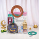 The pink-themed Imaan Play & Pray Kids Hamper neatly packed in a clear PVC carrying bag with round brown handles, styled with lit tea light candles, a golden crescent moon decoration, and a loose beaded tasbeeh resting on the table beside it.