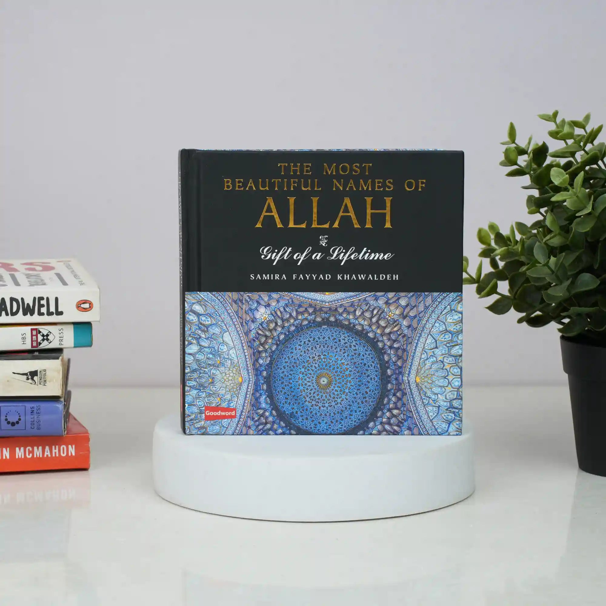 Book titled 'The Most Beautiful Names of Allah' on a white surface with books and a plant in the background. Main image