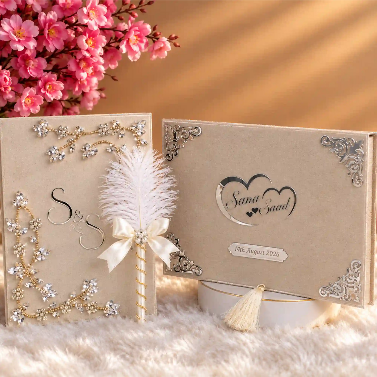 A styled wedding keepsake set featuring a cream-colored textured box with silver corner embellishments and a custom heart design, a matching decorated card with rhinestone corner accents, a white feather pen, and a cream tassel, arranged on a white faux fur surface with pink flowers in the background.