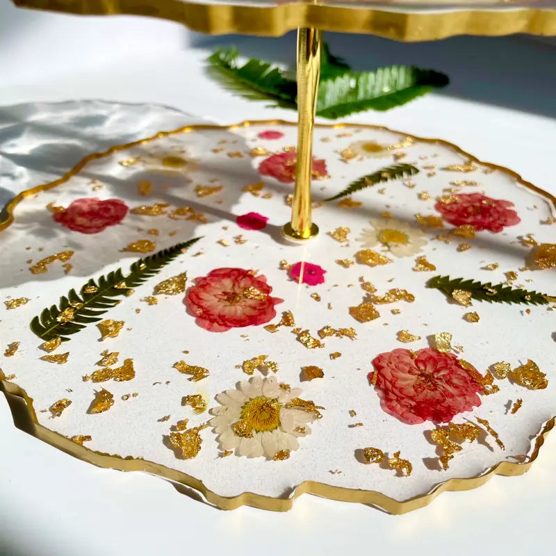 Two-tiered cake stand with floral and gold leaf design on a white surface.
