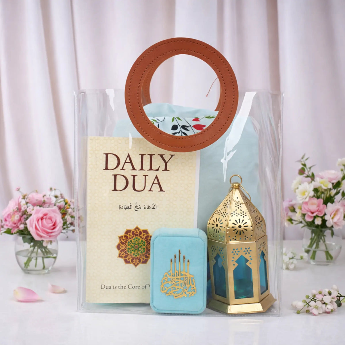 A clear PVC tote bag with round brown handles containing the Grace & Glow Women's Hamper. Inside the transparent bag, a Daily Dua book, a light blue velvet tasbeeh box with gold Arabic calligraphy, a gold metal lantern with blue glass, and a folded light blue prayer makhna are visible. The bag sits on a white surface flanked by small vases of pink and white flowers.
