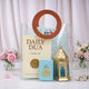 A clear PVC tote bag with round brown handles containing the Grace & Glow Women's Hamper. Inside the transparent bag, a Daily Dua book, a light blue velvet tasbeeh box with gold Arabic calligraphy, a gold metal lantern with blue glass, and a folded light blue prayer makhna are visible. The bag sits on a white surface flanked by small vases of pink and white flowers.