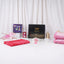 Front display of the Young Faith Blossoms Hamper items arranged on a white surface against a white curtain background. The collection features a closed black Hadiyah gift box with gold lettering, a folded pink prayer scarf, a pink rehal (book stand), a "Ramadan and Eid" children's book, a pink digital tasbeeh on a display card, a pink 33-bead tasbeeh, a mini golden lantern, and a folded plush pink kids janamaz.