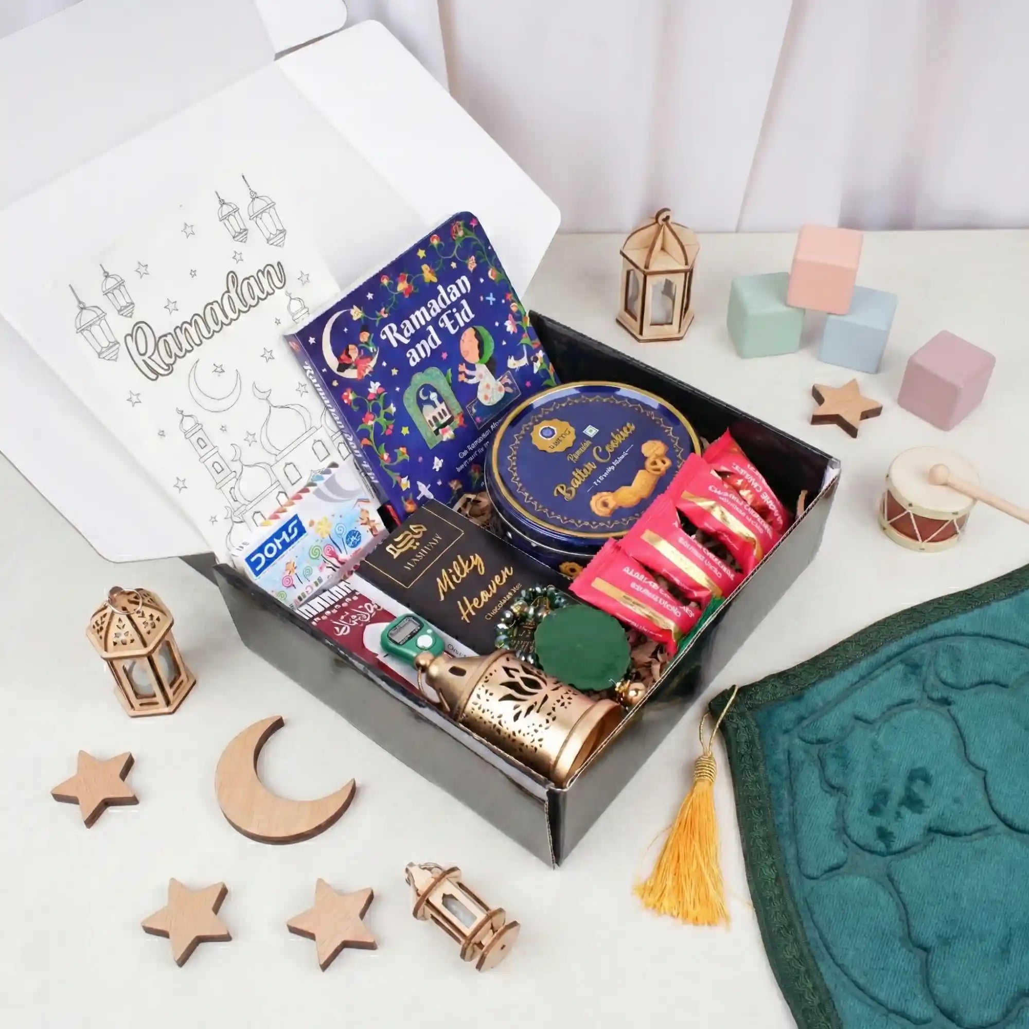 A top-down view of an open gift box filled with children's Islamic books, a cookie tin, chocolates, a green tasbeeh, and a mini golden lantern, surrounded by wooden moon and star decorations, building blocks, and a green velvet prayer mat. Main image