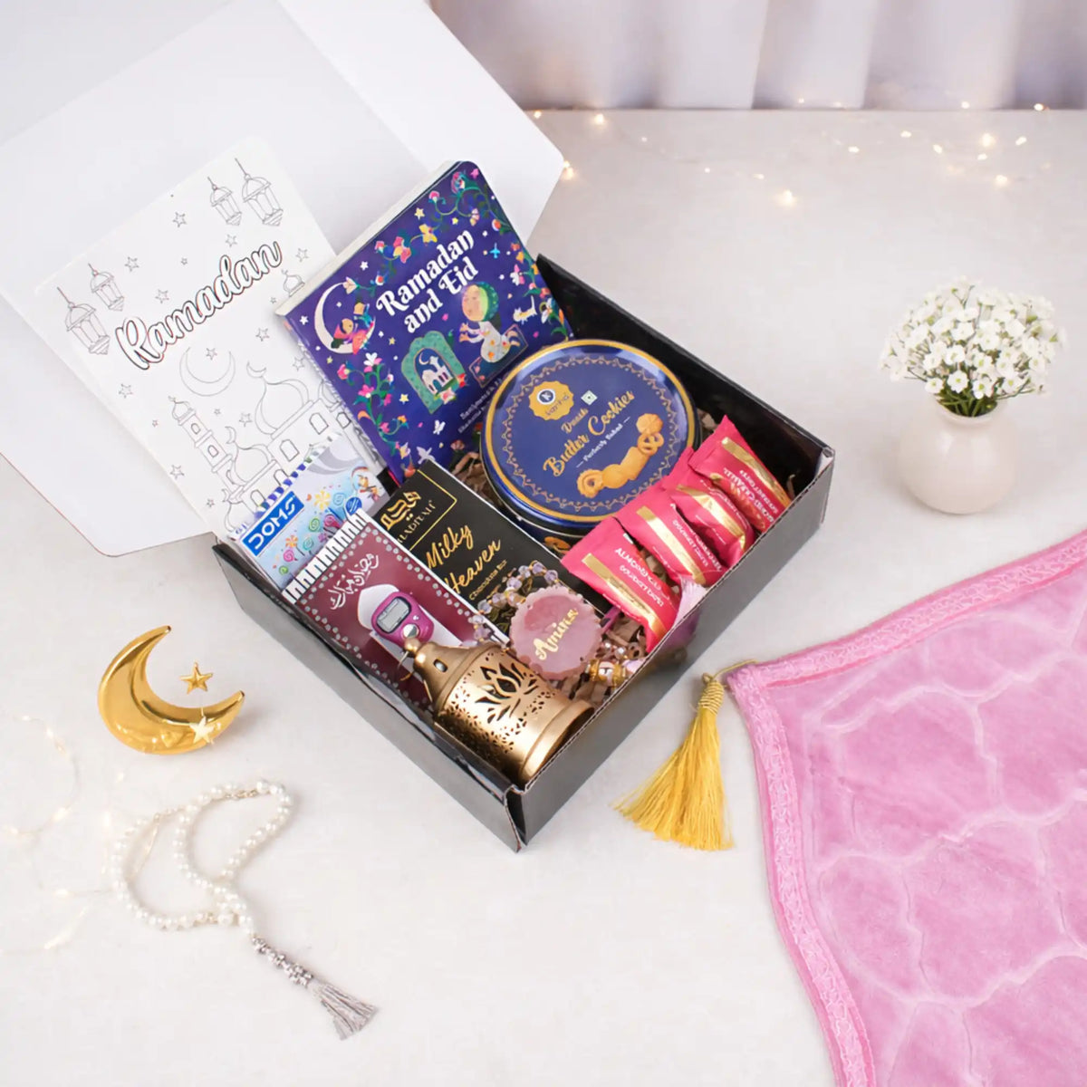 A top-down view of an open gift box containing a Ramadan and Eid book, crayons, a blue cookie tin, chocolates, a digital tasbeeh, and a mini golden lantern, sitting next to a folded pink velvet prayer mat and a white pearl tasbeeh.