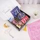 A top-down view of an open gift box containing a Ramadan and Eid book, crayons, a blue cookie tin, chocolates, a digital tasbeeh, and a mini golden lantern, sitting next to a folded pink velvet prayer mat and a white pearl tasbeeh.