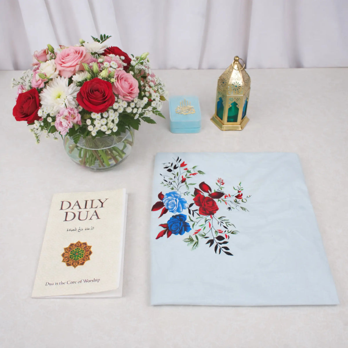 An overhead flat-lay view of the Grace & Glow Women's Hamper items arranged on a white surface. The layout includes a Daily Dua book, a neatly folded light blue prayer makhna featuring a vibrant red and blue rose print, a light blue velvet tasbeeh box, and a decorative gold metal lantern. A glass vase filled with red, pink, and white flowers sits in the top left corner.