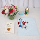 An overhead flat-lay view of the Grace & Glow Women's Hamper items arranged on a white surface. The layout includes a Daily Dua book, a neatly folded light blue prayer makhna featuring a vibrant red and blue rose print, a light blue velvet tasbeeh box, and a decorative gold metal lantern. A glass vase filled with red, pink, and white flowers sits in the top left corner.