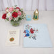 An overhead flat-lay view of the Grace & Glow Women's Hamper items arranged on a white surface. The layout includes a Daily Dua book, a neatly folded light blue prayer makhna featuring a vibrant red and blue rose print, a light blue velvet tasbeeh box, and a decorative gold metal lantern. A glass vase filled with red, pink, and white flowers sits in the top left corner.