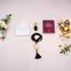 Top-down view of the Imaan Prestige Men's Hamper items laid flat on a white surface. The set features a white prayer cap, a small black attar bottle with a gold cap, a maroon Fortress of the Muslim dua book, and black crystal prayer beads with a gold medallion and tassel. The arrangement is framed by white roses, greenery, and a small lit golden lantern.