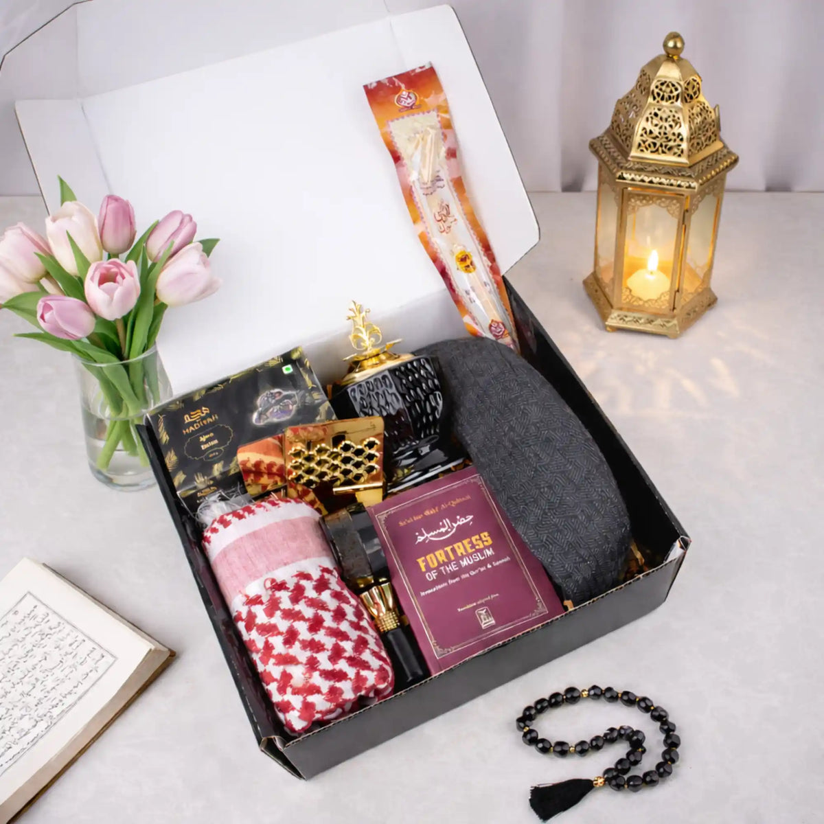 Top-down view of the open Noble Ibadah Men's Hamper box neatly packed with premium Islamic gifts, styled with pink tulips, an illuminated lantern, and black prayer beads.