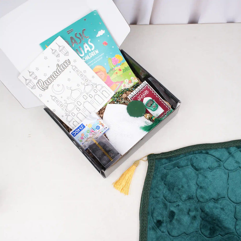 An overhead view of the Young Ibadah Kids Hamper for Boys. An open black gift box is filled with children's Ramadan essentials, including a Dua book, coloring sheets, crayons, packaged dates, a white prayer cap, and two tasbeehs. Next to the box, a dark green plush prayer mat with a golden tassel is partially unfolded on a white surface. Main image