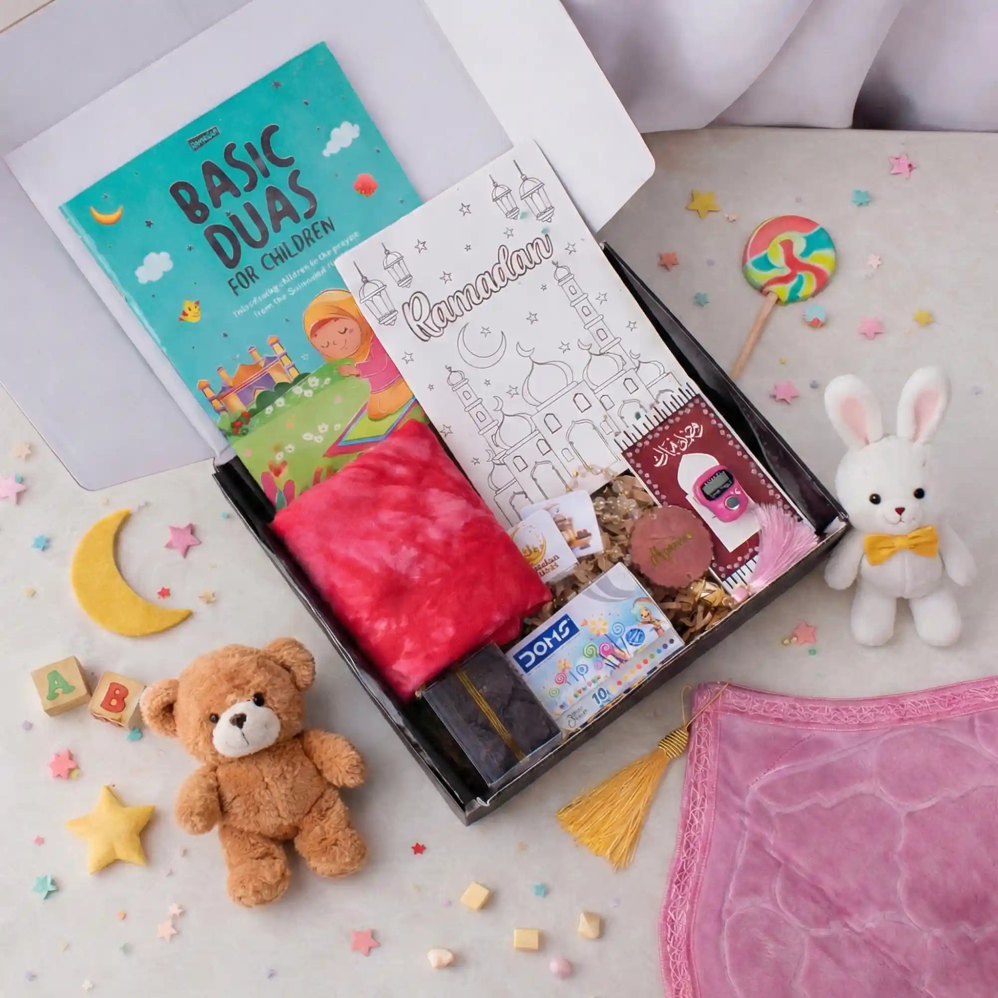 A flat-lay, top-down view of the Young Noorani Princess Hamper. An open black box is filled with a dua book, a coloring book, a red scarf, crayons, dates, and a pink digital tasbeeh. The box is surrounded by decorative props on a white surface, including a corner of a pink prayer mat with a gold tassel, a small brown teddy bear, a white stuffed bunny, wooden alphabet blocks, a colorful lollipop, and scattered pastel star and moon decorations. Main image