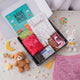 A flat-lay, top-down view of the Young Noorani Princess Hamper. An open black box is filled with a dua book, a coloring book, a red scarf, crayons, dates, and a pink digital tasbeeh. The box is surrounded by decorative props on a white surface, including a corner of a pink prayer mat with a gold tassel, a small brown teddy bear, a white stuffed bunny, wooden alphabet blocks, a colorful lollipop, and scattered pastel star and moon decorations.