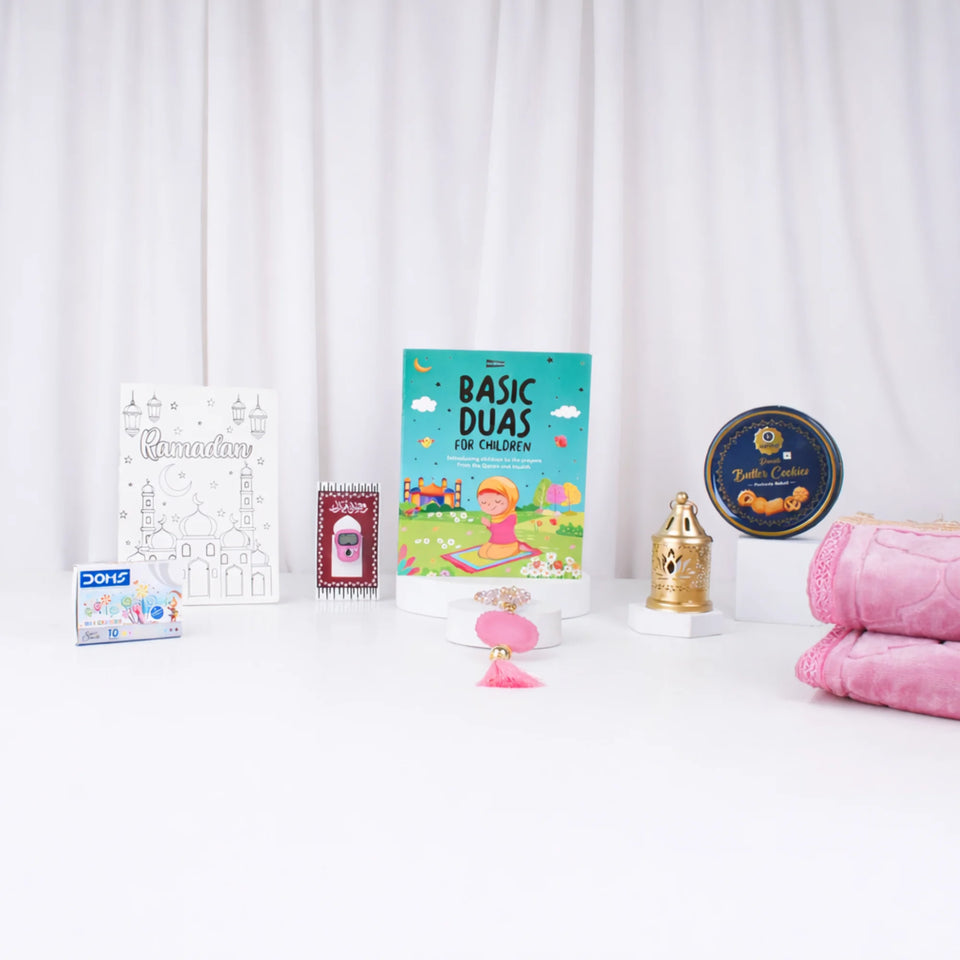 Front-facing display of the pink-themed kids hamper items arranged on a white surface, featuring a folded pink plush prayer mat, Basic Duas book, pink beaded and digital tasbeehs, crayons, coloring sheet, golden lantern, and butter cookie tin against a draped white curtain background.