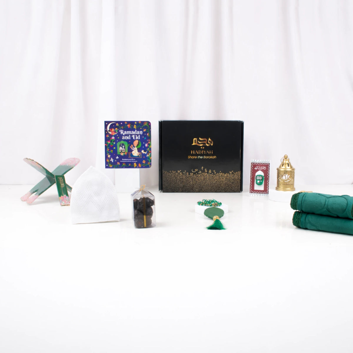 A straight-on view of the Little Learners of Deen Hamper contents arranged on a white surface next to a closed black gift box with gold lettering. The displayed items include a green painted wooden book stand (rehal), a white prayer cap (topi), a "Ramadan and Eid" children's book, a clear package of dates, a green beaded tasbeeh, a digital tasbeeh mounted on a decorative card, a golden mini lantern, and a folded green prayer mat.