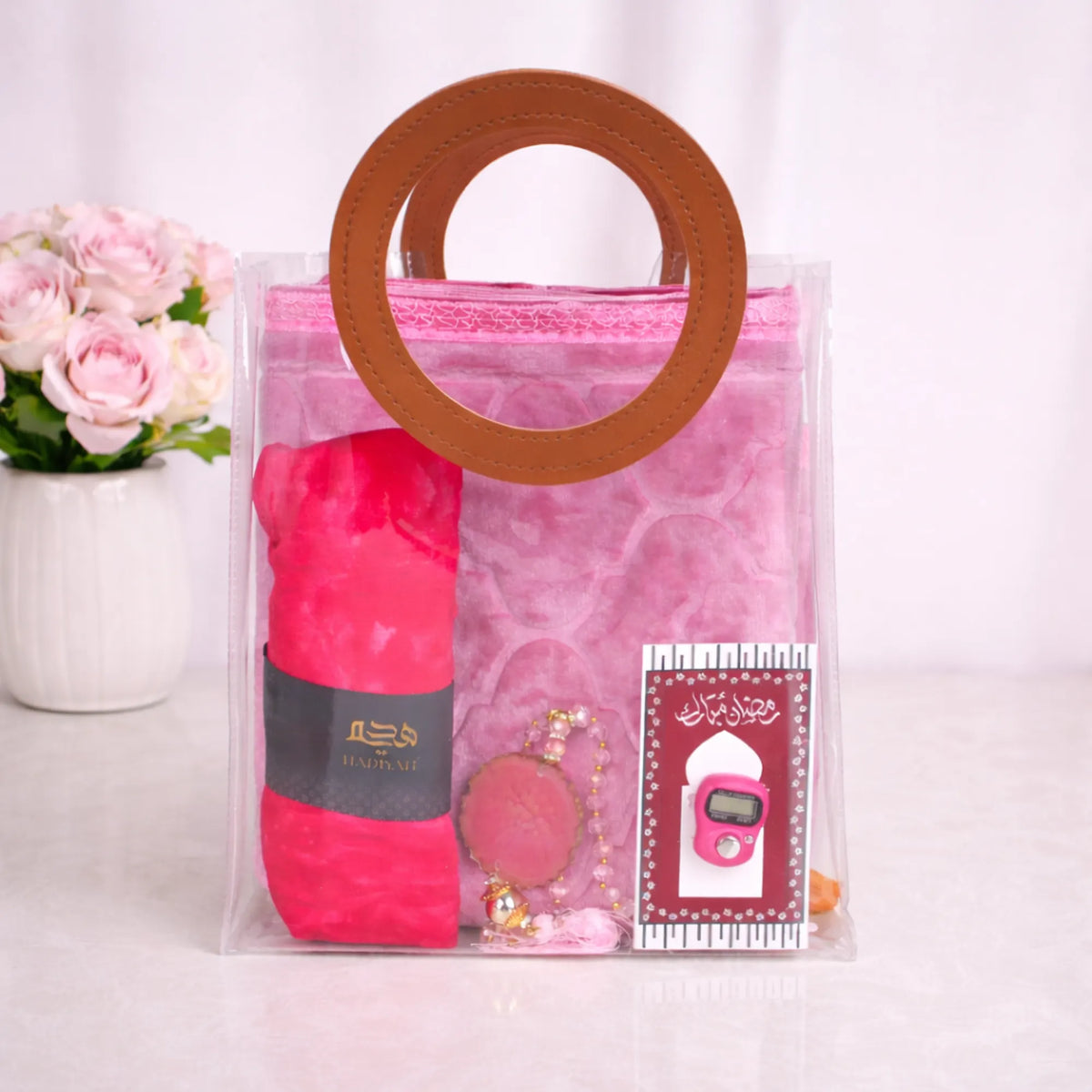 Front view of the pink Little Soul Essentials Hamper packaged in a clear PVC tote bag with brown round handles. Visible inside are a folded light pink kids' janamaz, a rolled bright pink prayer makhana, a pink beaded tasbeeh with an agate stone, and a digital tasbeeh mounted on a decorative card.