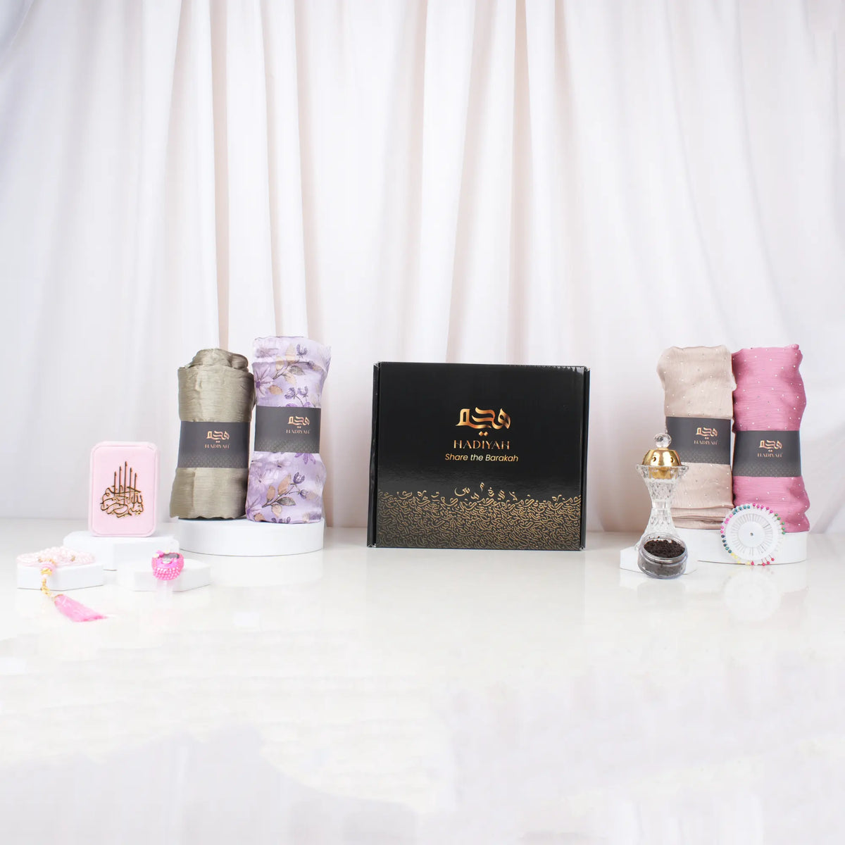 A front-facing display featuring a closed black Hadiyah gift box adorned with elegant gold calligraphy and the text "Share the Barakah." Displayed upright on white pedestals around the box are four rolled chiffon hijabs in olive green, purple floral, textured beige, and solid pink. The arrangement also includes a pink velvet box with gold detailing, a beaded pink tasbeeh, a pink digital tally counter, a crystal bakhoor burner, a small jar of bakhoor, and a hijab pin wheel, all set against a soft, draped whi