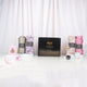 A front-facing display featuring a closed black Hadiyah gift box adorned with elegant gold calligraphy and the text "Share the Barakah." Displayed upright on white pedestals around the box are four rolled chiffon hijabs in olive green, purple floral, textured beige, and solid pink. The arrangement also includes a pink velvet box with gold detailing, a beaded pink tasbeeh, a pink digital tally counter, a crystal bakhoor burner, a small jar of bakhoor, and a hijab pin wheel, all set against a soft, draped whi