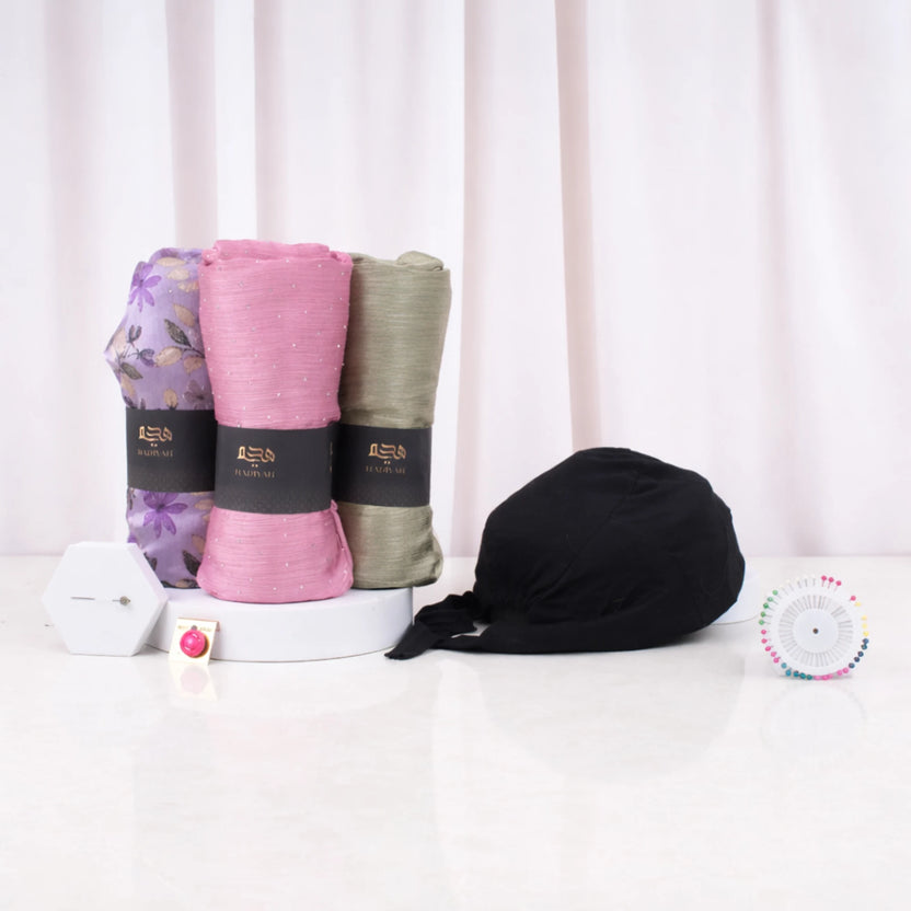 Front display of the Modest Style Set Women's Hamper contents on a white surface. Three rolled hijabs in purple floral, pink, and olive green stand upright on a small display riser, next to a black hijab cap, a diamond pin, a hijab magnet, and a wheel of colorful pins. Main image