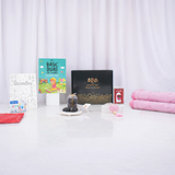A front-facing display of the Young Noorani Princess Hamper items arranged on a white surface against a white draped curtain. The collection includes a black "Hadiyah" gift box in the center, flanked by a rolled pink plush prayer mat, a "Basic Duas for Children" book, a Ramadan coloring book, a box of DOMS crayons, a small package of dates, a red scarf, and a pink digital tasbeeh.