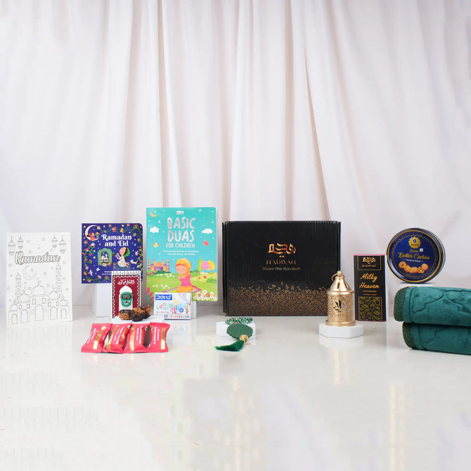 A front-facing arrangement of the green-themed Ultimate Kids Celebration Hamper, displaying a black Hadiyah gift box, a folded green velvet prayer mat, children's Islamic books, crayons, a cookie tin, chocolates, a green tasbeeh, and a mini golden lantern.