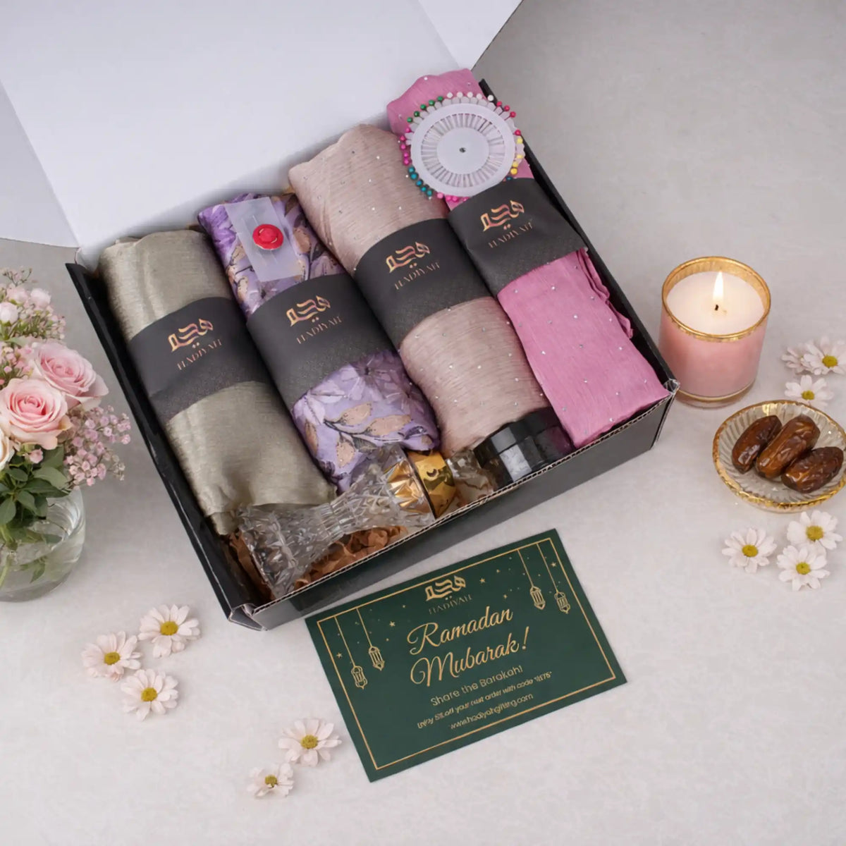 An overhead view of an open black gift box containing four elegantly rolled chiffon hijabs in olive green, purple floral, textured beige, and solid pink, each wrapped with a branded black band. Inside the box rests a crystal bakhoor burner, a small dark jar, and a colorful wheel of hijab pins. A dark green "Ramadan Mubarak!" greeting card sits in front of the box, beautifully styled with fresh pink roses, scattered white daisies, a lit pink candle, and a small decorative plate of dates.