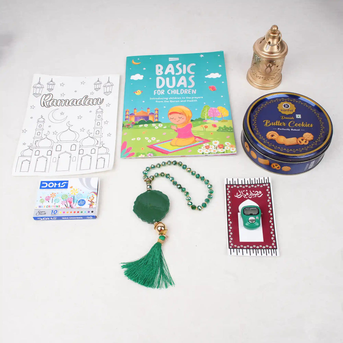 Top-down flat lay of the green-themed Imaan Play & Pray Kids Hamper contents, displaying a Basic Duas for Children book, a Ramadan coloring sheet, crayons, a green digital tasbeeh, a green 33-bead tasbeeh with a tassel, a mini golden lantern, and a blue tin of Danish Butter Cookies.