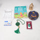 Top-down flat lay of the green-themed Imaan Play & Pray Kids Hamper contents, displaying a Basic Duas for Children book, a Ramadan coloring sheet, crayons, a green digital tasbeeh, a green 33-bead tasbeeh with a tassel, a mini golden lantern, and a blue tin of Danish Butter Cookies.