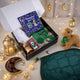 An overhead view of an open gift box filled with children's Islamic items, including a "Ramadan and Eid" book, a white prayer cap, and prayer beads. The box is surrounded by a festive, warmly lit setting featuring fairy lights, a lit golden lantern, a crescent moon-shaped dish filled with dates, a small plush camel wearing a green ribbon, and a soft green quilted prayer mat laid out beside it.