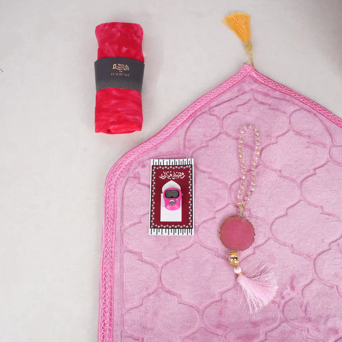 Top-down view of the Little Soul Essentials Hamper contents laid flat on a light surface. It features a light pink quilted kids' prayer mat, a pink digital tasbeeh on a printed card, a matching pink 33-bead tasbeeh with a large agate stone and tassel, and a bright pink rolled prayer makhana secured with a black Hadiyah band.