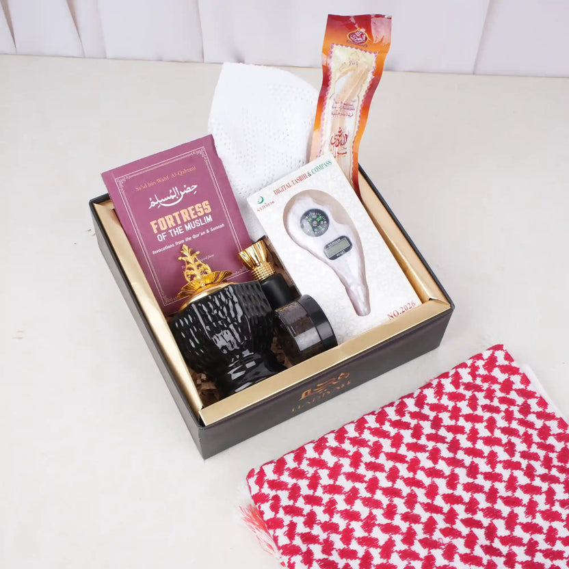 An overhead view of the Son of the Ummah Men's Gift Hamper. A black gift box with gold inner lining contains a white prayer cap, a packaged miswak, a maroon 'Fortress of the Muslim' book, a boxed digital tasbeeh with a compass, a black and gold bakhoor burner, a small black jar of bakhoor, and an attar bottle nestled inside. A red and white checkered ghutra rests on the white surface outside the bottom right corner of the box. Main image